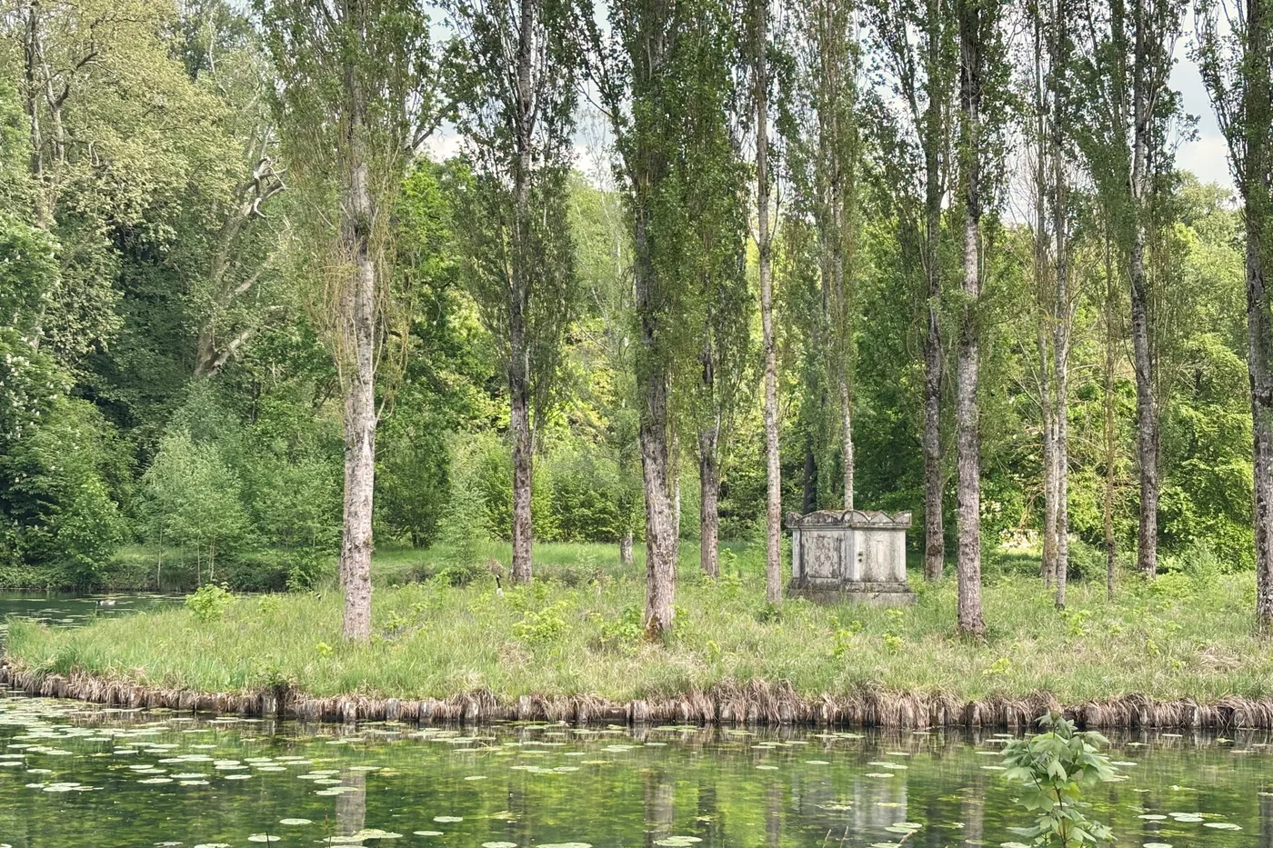 Un monument funéraire de pierre blanche se dresse sur une île plantée de grands peupliers, au-delà d’une eau couverte de nénuphars.