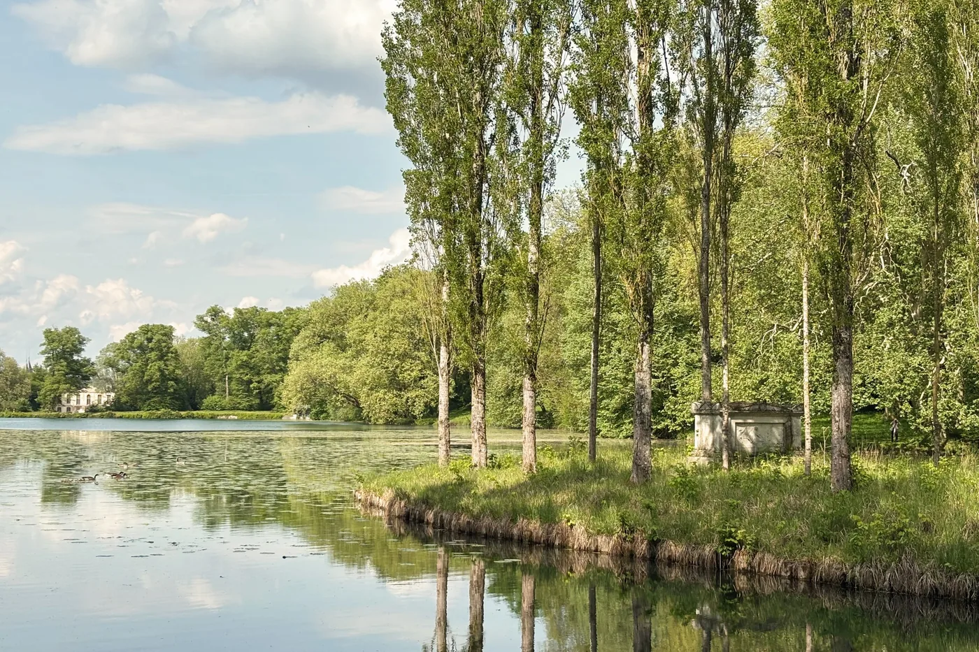 Un lac calme reflète une rangée de grands peupliers ; sur l'île, à droite, un monument de pierre blanche ; au loin, une demeure claire parmi les arbres.