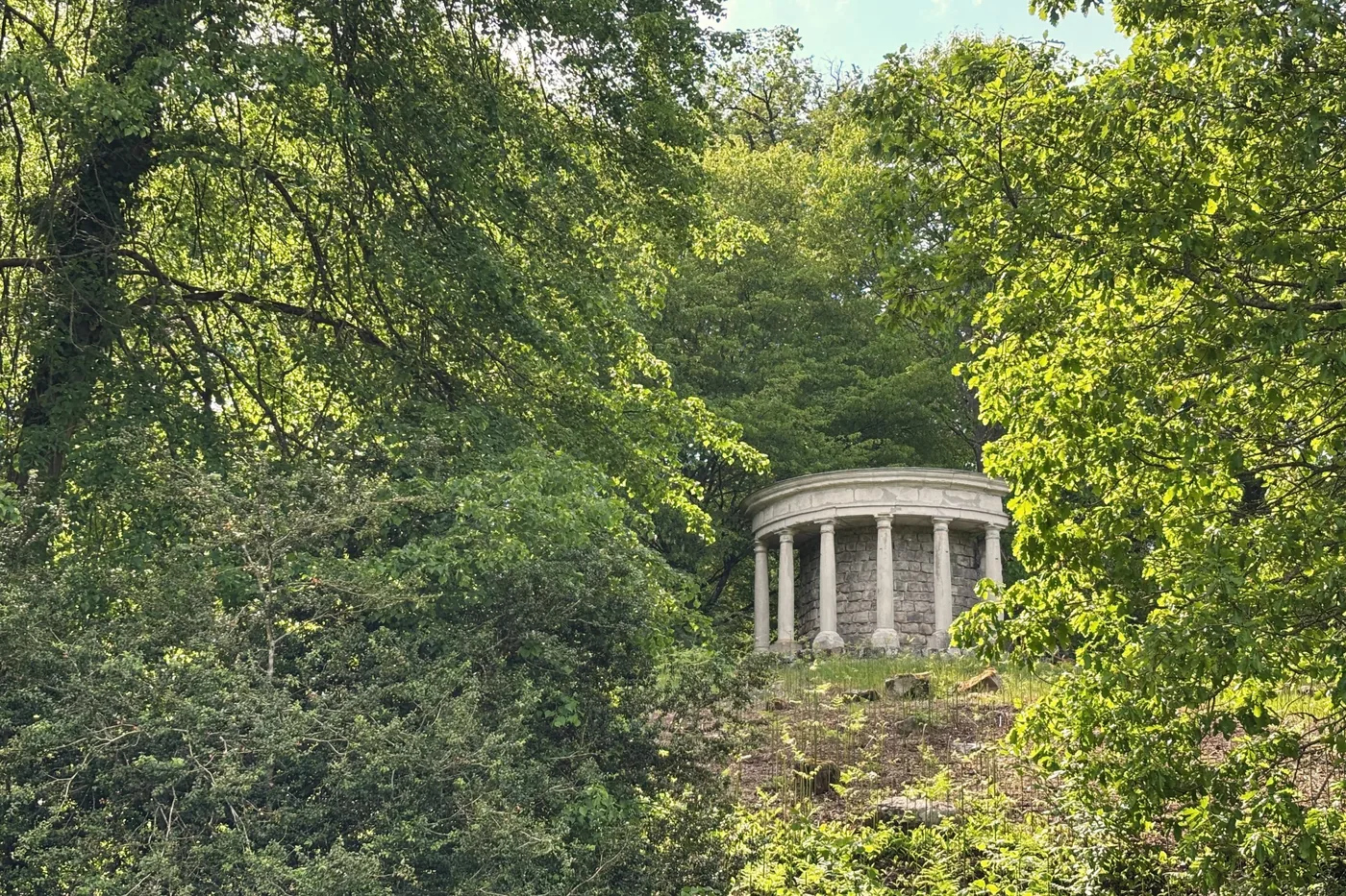 Une rotonde à colonnes, aux murs de pierres brutes, apparaît entre les feuillages en haut d’une pente.