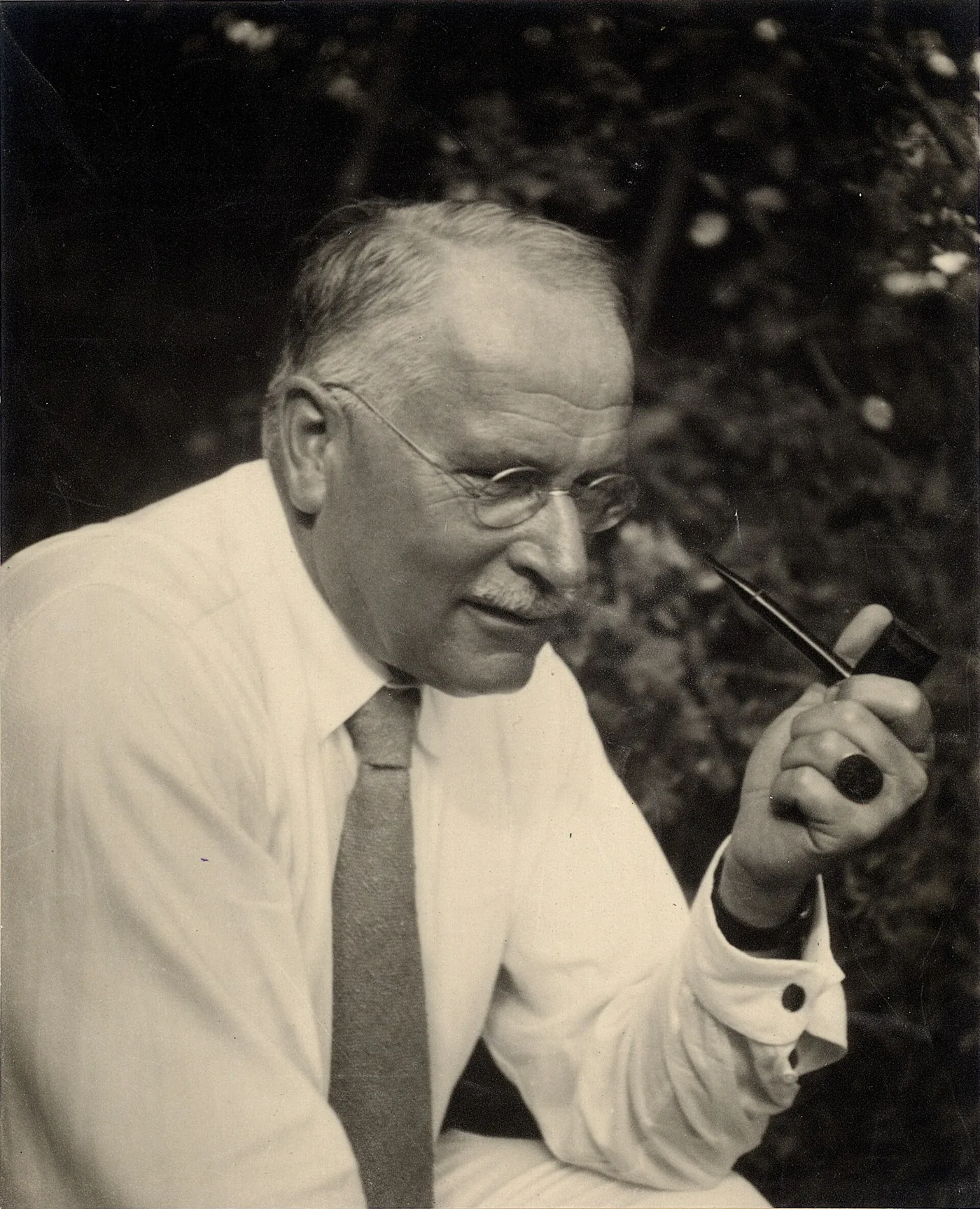 Photographie en noir et blanc d'un homme âgé à lunettes rondes et moustache blanche, en chemise claire et cravate, tenant une pipe, devant un feuillage sombre.