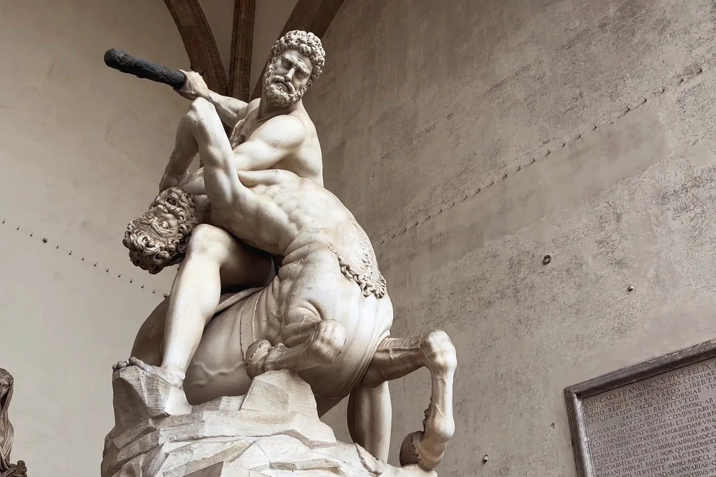 Groupe de marbre : un homme barbu brandit une massue et maîtrise un centaure dont il tient la tête renversée, devant un mur clair et une plaque gravée.