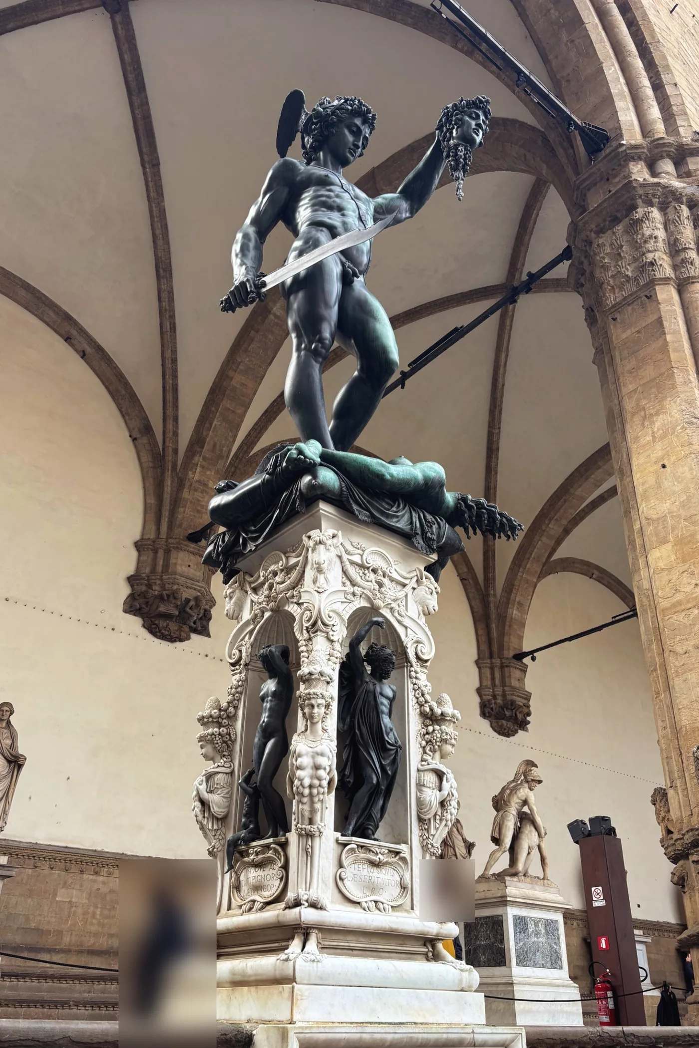 Statue de bronze d'un jeune homme nu au casque ailé, une épée dans une main et une tête coupée dans l'autre, debout sur un corps, au sommet d'un haut piédestal de marbre sculpté, sous des arcs gothiques.
