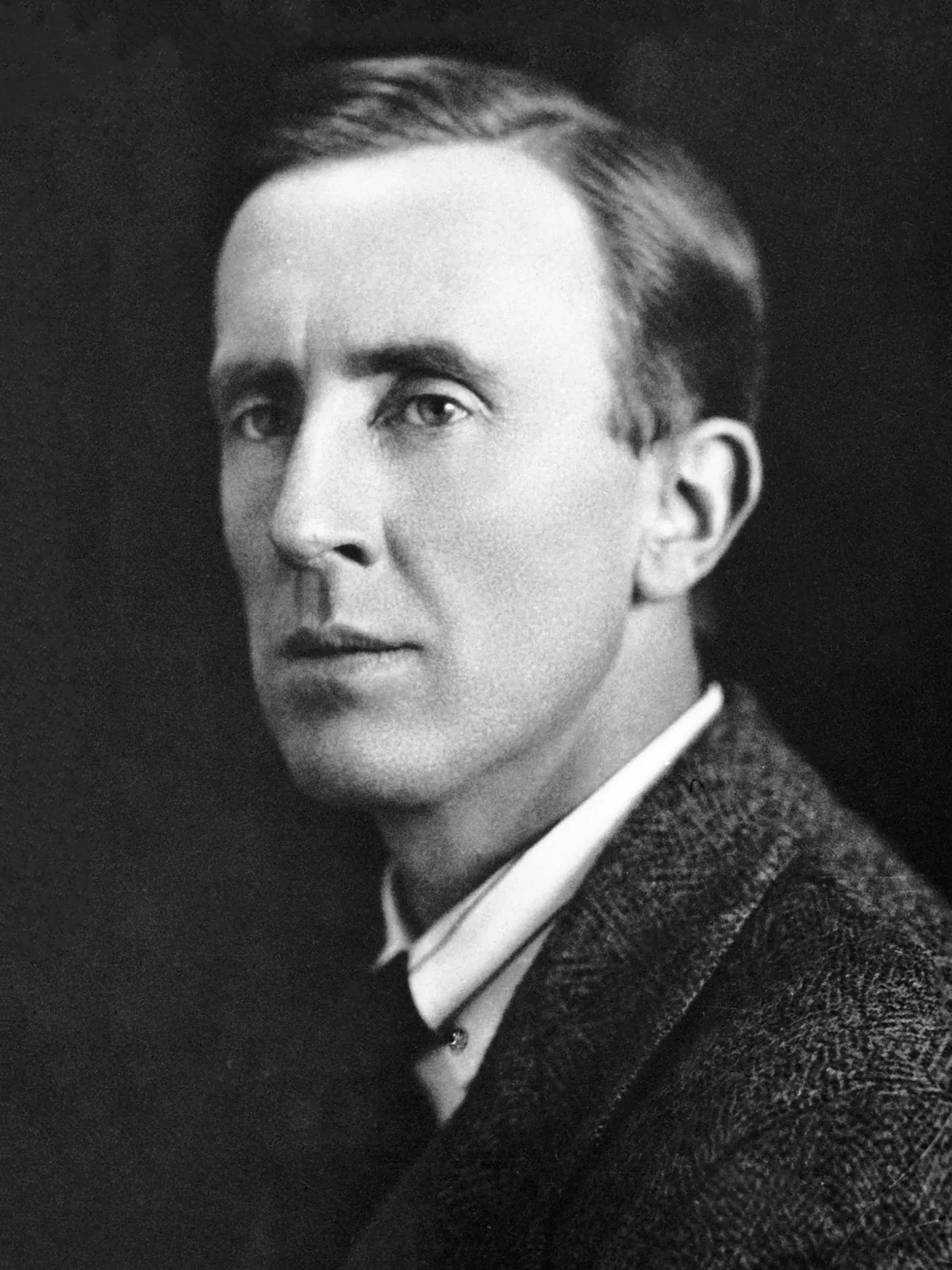 Portrait en noir et blanc d'un homme de trois quarts, cheveux coiffés en arrière, col blanc et veste de tweed, sur fond sombre.